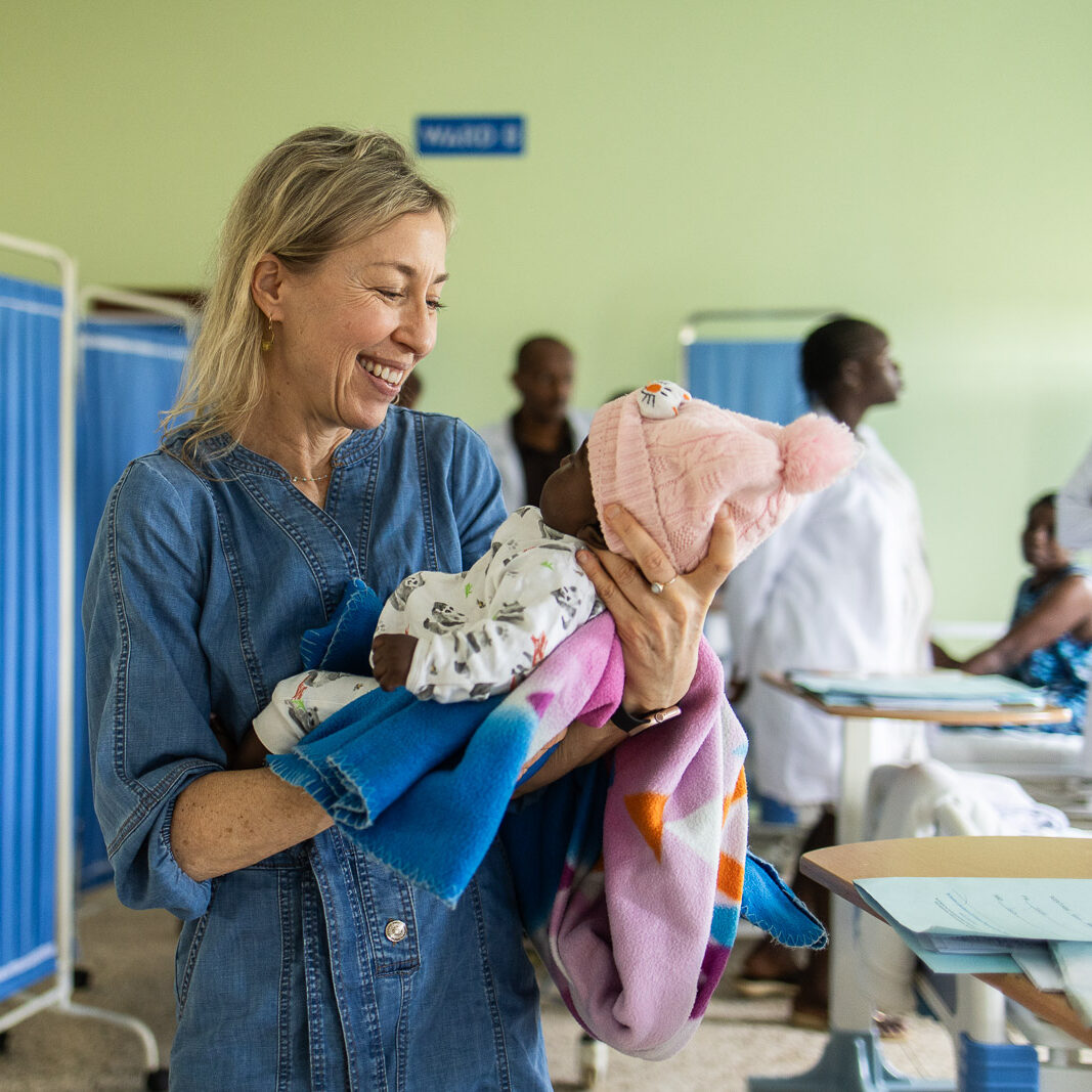 Bonnie Ruder in a denim dress cradles a baby wearing a pink knit hat inside a hospital ward in Uganda.