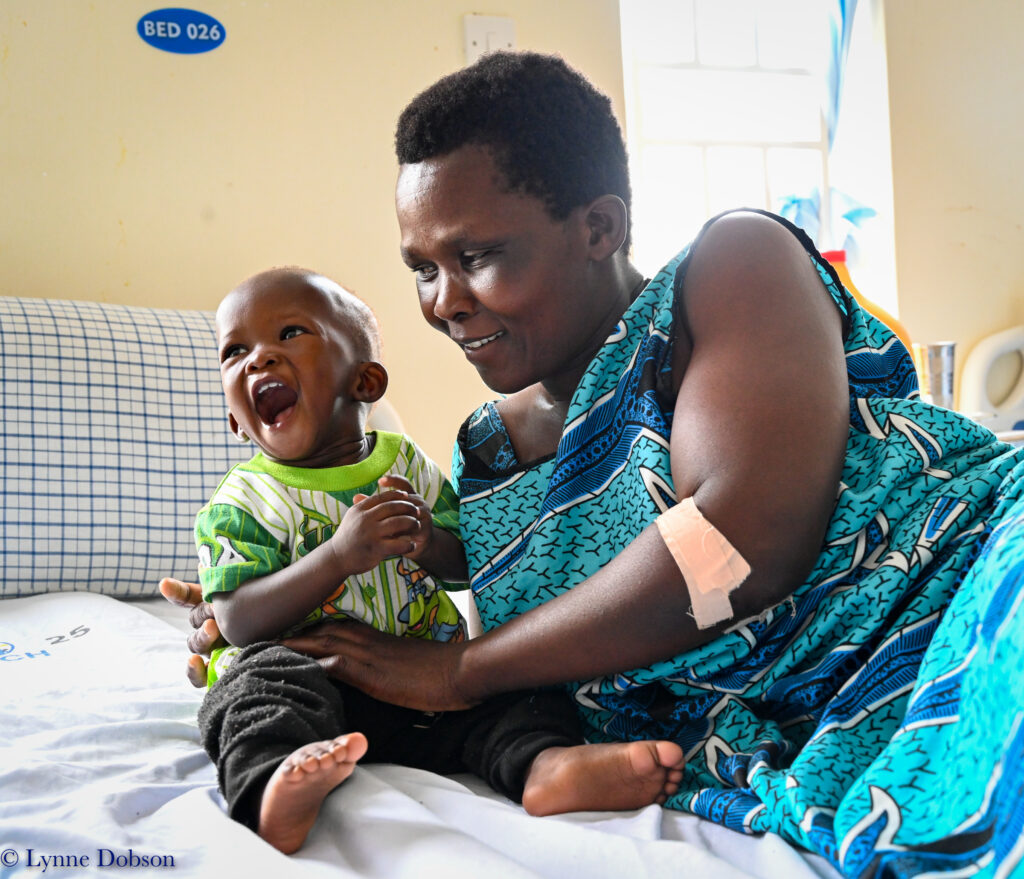 Terrewode_Uganda_2022-493 Smiling mother in a blue patterned dress holds a laughing toddler on a hospital bed at Terrewode Women's Community Hospital in Uganda.
