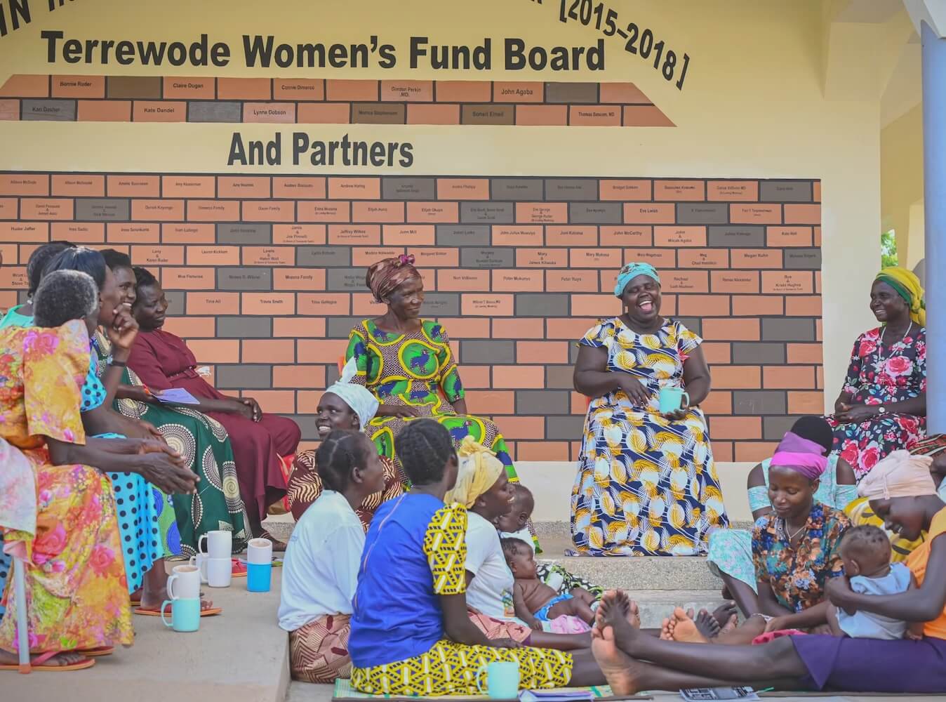 Women in colorful clothing share laughter and conversation while seated together with babies outside Terrewode Women’s Community Hospital in Soroti, Uganda, beneath a donor recognition wall honoring Terrewode Women’s Fund board members and partners.